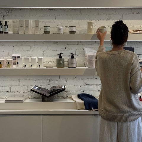 A woman viewed from behind stands in a retail space with white shelves displaying many bottles, jars, and boxes against a white brick wall.